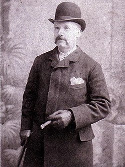 Lusk has a moustache and wears a bowler hat, topcoat and leather gloves. He holds a cane in the right hand and a cigar in the left.